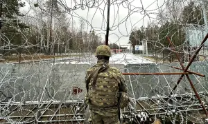 Żołnierz w mundurze obserwuje teren przez zasieki z drutu kolczastego. Widoczna jest granica polsko-białoruska, z budynkami i znakami po drugiej stronie.