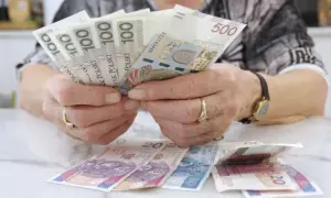 Dłonie z obrączkami trzymają banknoty 100 i 500 zł. Na stole leżą banknoty 20 zł. Czerwcowe emerytury już są!