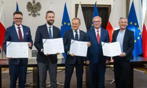 Pięciu mężczyzn w garniturach stoi przed flagami Polski i UE, trzymając dokumenty.