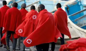Grupa migrantów w czerwonych kocach z białym krzyżem, po trudnej podróży, szuka schronienia.