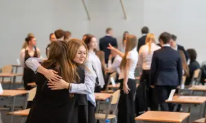 Dwie uczennice przytulają się, gdy wyniki matur są ogłaszane. W tle widać innych uczniów i sale lekcyjne.