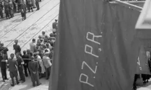 Tłum ludzi zgromadzony na ulicy, na pierwszym planie widoczna wielka, ciemna tkanina z napisem PZPR.