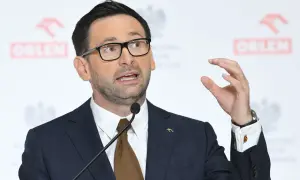 Mężczyzna w garniturze i okularach przemawia na tle logo Orlenu. Analiza fuzji Orlenu z Lotosem za rządów PiS.