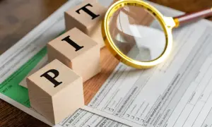 Drewniane klocki z literami "PIT" leżą na formularzu podatkowym. Złota lupa przybliża szczegóły. Czas zacząć myśleć, od kiedy można rozliczać PIT.