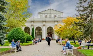 Ludzie siedzą na ławkach przed budynkiem, ciesząc się jesiennym parkiem. Dowiedz się o nowe zasady wyjazdu do sanatorium na NFZ.