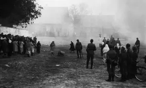 Czarno-białe zdjęcie przedstawia grupę ludzi, w tym żołnierzy, zgromadzonych na otwartym terenie przed budynkami.