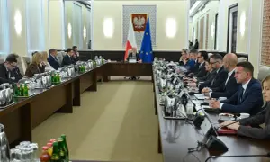 Uczestnicy spotkania przy długim stole, dyskutują o budżetowym deficycie. Na stole napoje i dokumenty.