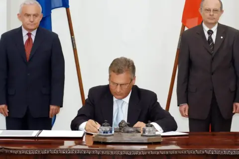 Uroczyste podpisanie dokumentów, symboliczny moment, kiedy Polska weszła do Unii. Trzech mężczyzn w garniturach, flagi Polski i UE.