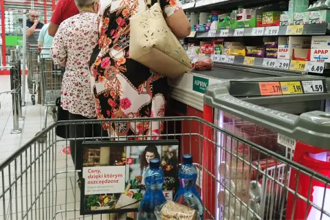 Kolejka w sklepie, ludzie robią zakupy. Widać ceny produktów, przypominając o zakazie handlu w niedziele i święta.