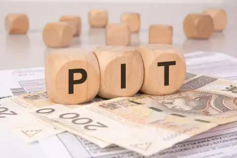 Drewniane klocki z literami "PIT" leżą na banknotach 200 zł i dokumentach podatkowych. To symboliczne przedstawienie rozliczenia podatku, gdzie darowizna może wpłynąć na Twój PIT.