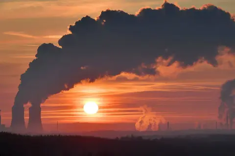 Zachód słońca nad przemysłowym krajobrazem, gdzie dymiące kominy kontrastują z pomarańczowym niebem. To obraz, który może symbolizować wyzwania związane z polityką klimatyczną i stosunkiem PiS do Zielonego Ładu.