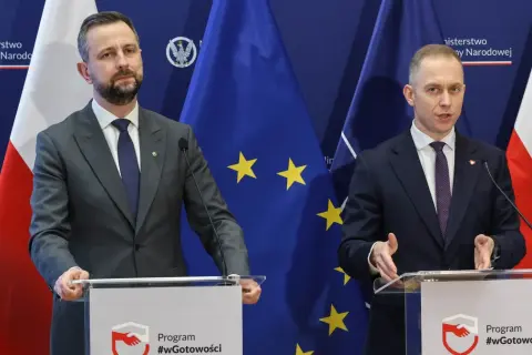 Dwóch mężczyzn w garniturach przemawia za mównicami. Tło stanowią flagi Polski i UE. Tematem dyskusji mogło być to, jak rządy PiS zmieniły strukturę polskiego wojska.