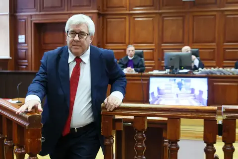 Mężczyzna w garniturze i krawacie wchodzi na salę sądową. Sędziowie w togach siedzą za stołem. Proces polityków PiS trwa.
