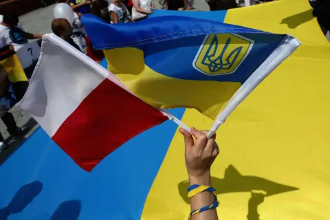 Dłoń trzyma polską i ukraińską flagę. W tle widać ludzi i wielką ukraińską flagę. To symboliczne przedstawienie wpływu koalicji obywatelskiej na relacje Polski z Ukrainą.
