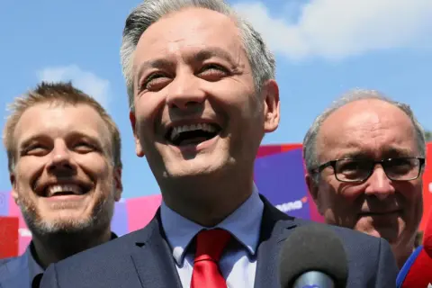 Uśmiechnięty polityk z czerwonym krawatem, w otoczeniu innych mężczyzn. Analiza struktury elektoratu partii, kogo reprezentuje lewica.