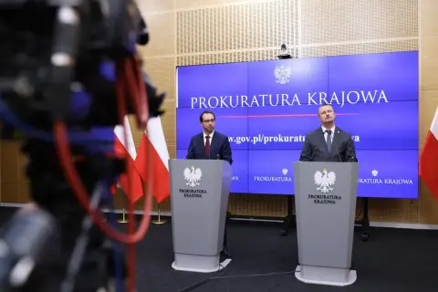 Dwóch mężczyzn w garniturach stoi za mównicami z godłem Polski, na tle ekranu z napisem "Prokuratura Krajowa".