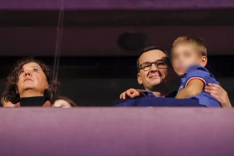 Mateusz Morawiecki z dziećmi, uśmiechnięty, trzyma na rękach jedno z dzieci. Obok kobieta patrzy w górę.