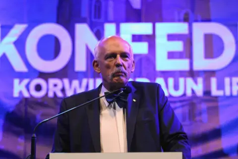 Janusz Korwin-Mikke, znany z kontrowersyjnych poglądów, przemawia na tle napisu "Konfederacja".