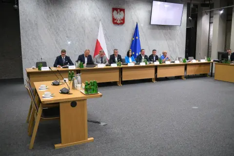 Osiem osób siedzi przy stole w sali konferencyjnej, przed nimi flagi Polski i UE.