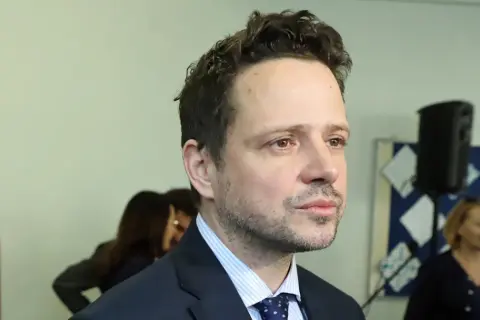 Rafał Trzaskowski, którego wykształcenie budzi zainteresowanie, w garniturze i krawacie. W tle inne osoby i tablica.