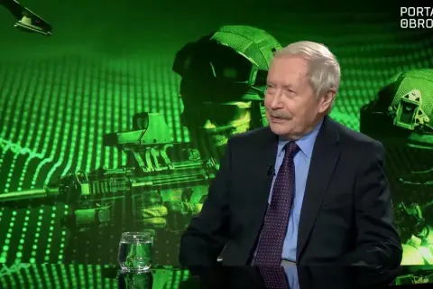 Starszy mężczyzna w garniturze rozmawia na tle zielonej grafiki z żołnierzami i bronią. To może być dyskusja o układzie warszawskim.