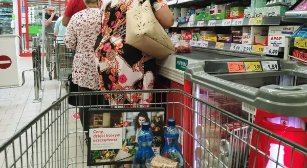 Kolejka w sklepie, ludzie robią zakupy. Widać ceny produktów, przypominając o zakazie handlu w niedziele i święta.