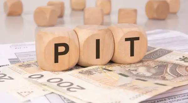 Drewniane klocki z literami "PIT" leżą na banknotach 200 zł i dokumentach podatkowych. To symboliczne przedstawienie rozliczenia podatku, gdzie darowizna może wpłynąć na Twój PIT.