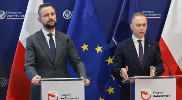 Dwóch mężczyzn w garniturach przemawia za mównicami. Tło stanowią flagi Polski i UE. Tematem dyskusji mogło być to, jak rządy PiS zmieniły strukturę polskiego wojska.
