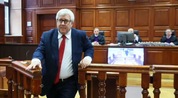 Mężczyzna w garniturze i krawacie wchodzi na salę sądową. Sędziowie w togach siedzą za stołem. Proces polityków PiS trwa.
