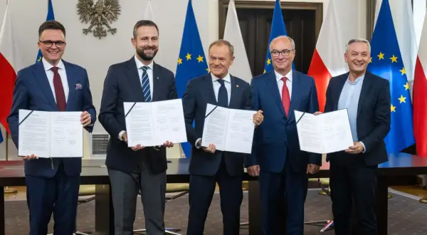 Pięciu mężczyzn w garniturach stoi przed flagami Polski i UE, trzymając dokumenty.