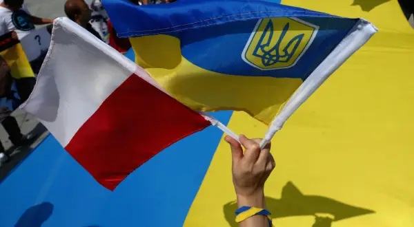 Dłoń trzyma polską i ukraińską flagę. W tle widać ludzi i wielką ukraińską flagę. To symboliczne przedstawienie wpływu koalicji obywatelskiej na relacje Polski z Ukrainą.