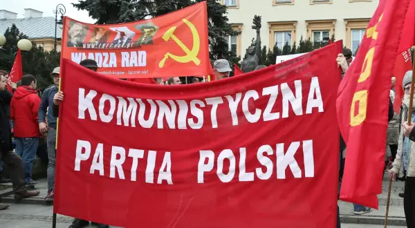 Ludzie niosą czerwony transparent "KOMUNISTYCZNA PARTIA POLSKI" i flagę z sierpem i młotem. To manifestacja poparcia dla socjalizmu.