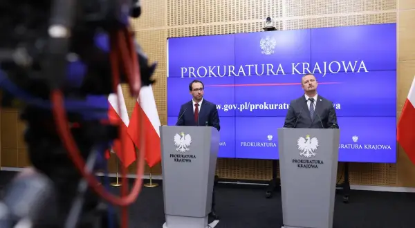 Dwóch mężczyzn w garniturach stoi za mównicami z godłem Polski, na tle ekranu z napisem "Prokuratura Krajowa".