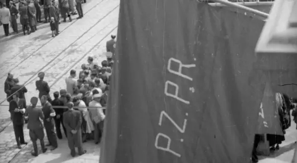 Tłum ludzi zgromadzony na ulicy, na pierwszym planie widoczna wielka, ciemna tkanina z napisem PZPR.