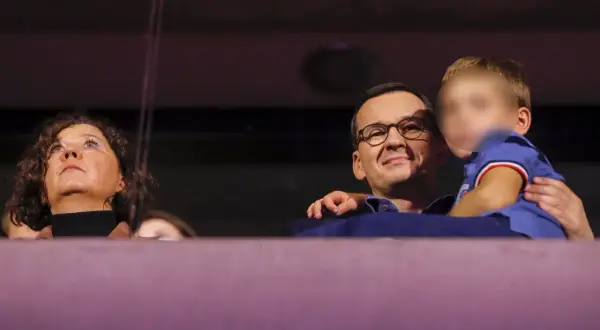Mateusz Morawiecki z dziećmi, uśmiechnięty, trzyma na rękach jedno z dzieci. Obok kobieta patrzy w górę.