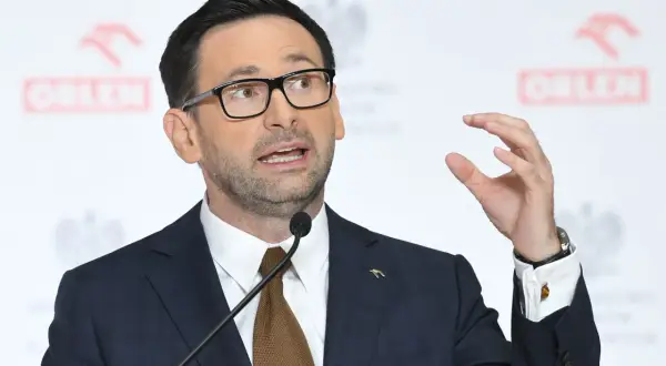 Mężczyzna w garniturze i okularach przemawia na tle logo Orlenu. Analiza fuzji Orlenu z Lotosem za rządów PiS.