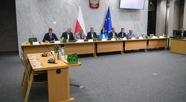Osiem osób siedzi przy stole w sali konferencyjnej, przed nimi flagi Polski i UE.