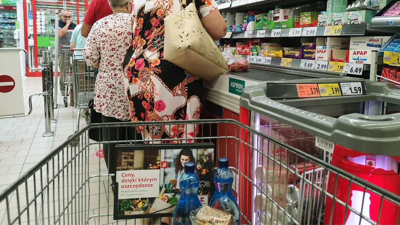 Kolejka w sklepie, ludzie robią zakupy. Widać ceny produktów, przypominając o zakazie handlu w niedziele i święta.