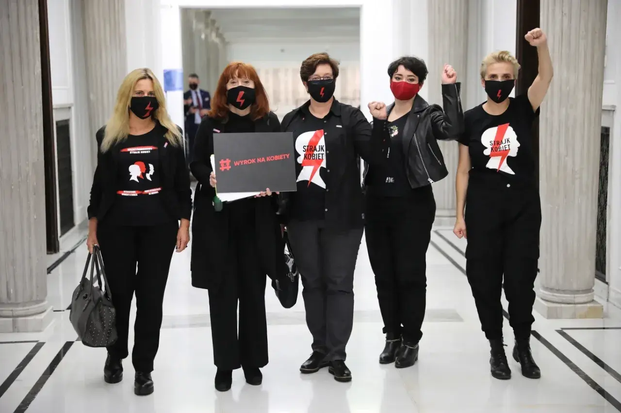 Kobiety w czarnych koszulkach "Strajk Kobiet" i maskach, z hasłem "Wyrok na kobiety", symbolizują siłę lewicy i jej zaangażowanie w protesty.