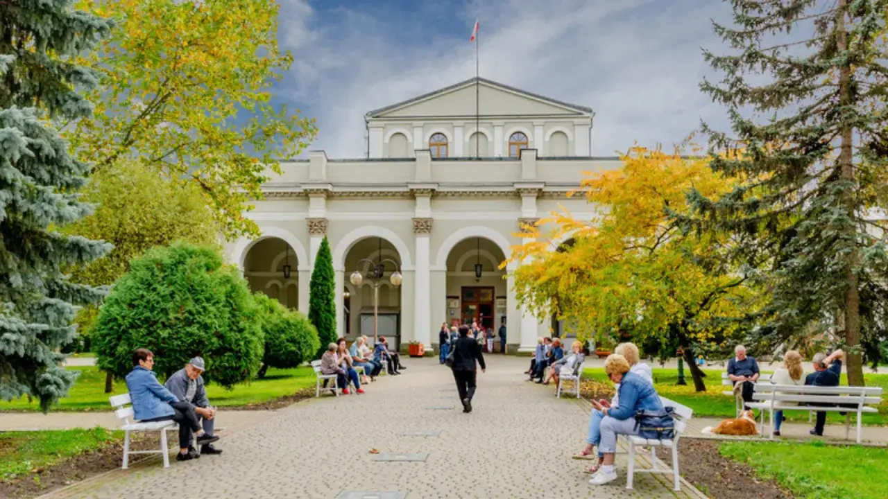 Ludzie siedzą na ławkach przed budynkiem, ciesząc się jesiennym parkiem. Dowiedz się o nowe zasady wyjazdu do sanatorium na NFZ.