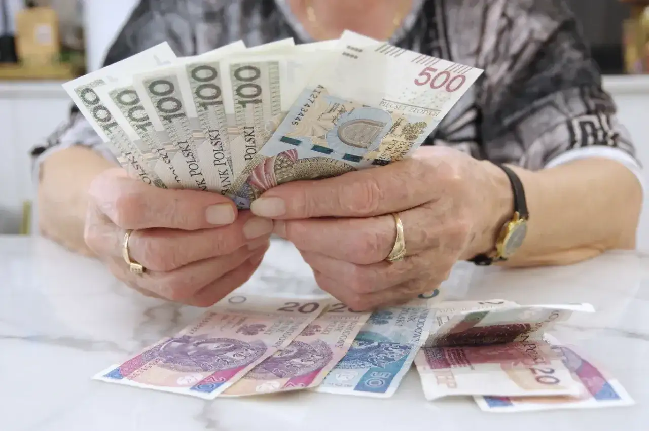 Dłonie z obrączkami trzymają banknoty 100 i 500 zł. Na stole leżą banknoty 20 zł. Czerwcowe emerytury już są!