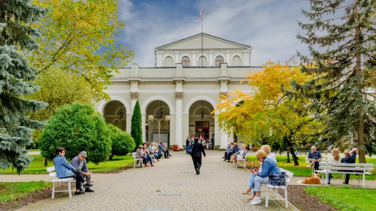 Ludzie siedzą na ławkach przed budynkiem, ciesząc się jesiennym parkiem. Dowiedz się o nowe zasady wyjazdu do sanatorium na NFZ.