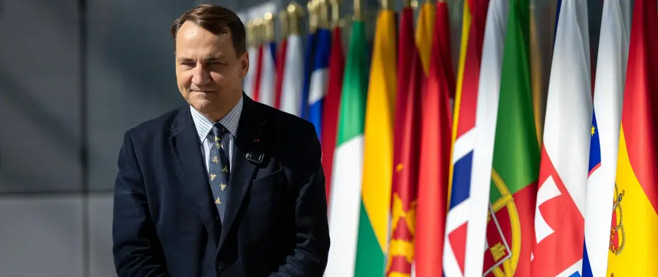 Radosław Sikorski i dyplomacja w wizji Koalicji Obywatelskiej. Polityk na tle flag państwowych, symbolizujących międzynarodowe relacje.