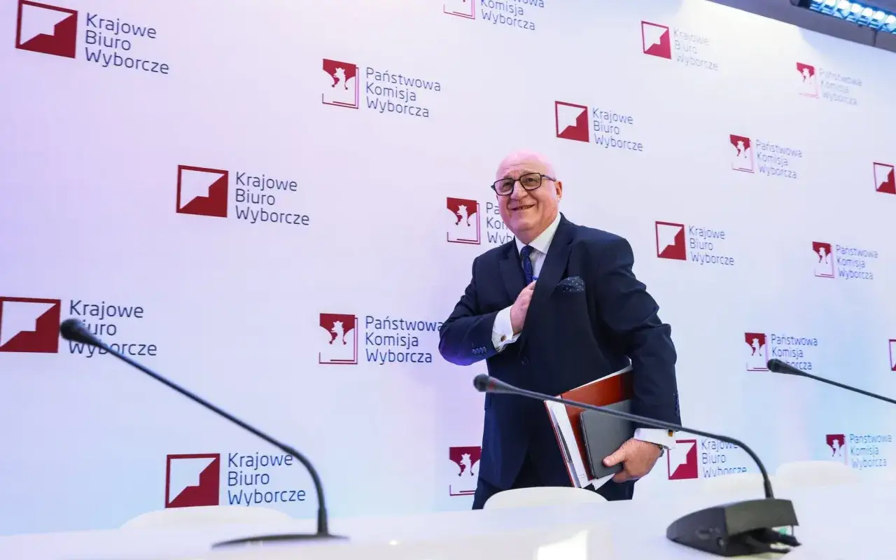 Mężczyzna w garniturze, trzymający dokumenty, uśmiecha się na tle logo PKW. Czy to powody odrzucenia sprawozdania o subwencji dla PiS?