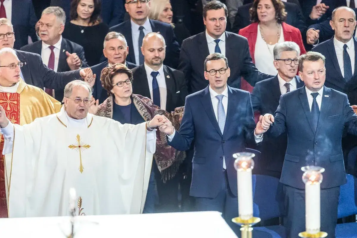 Premier Morawiecki i inni politycy PiS trzymają się za ręce z księdzem. W tle widać ludzi, którzy mogą być beneficjentami dotacji dla Ojca Rydzyka.