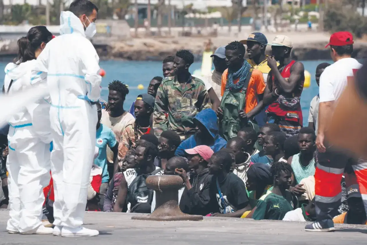 Grupa młodych mężczyzn, migrantów, stoi na nabrzeżu. Obok nich pracownicy w białych kombinezonach ochronnych.