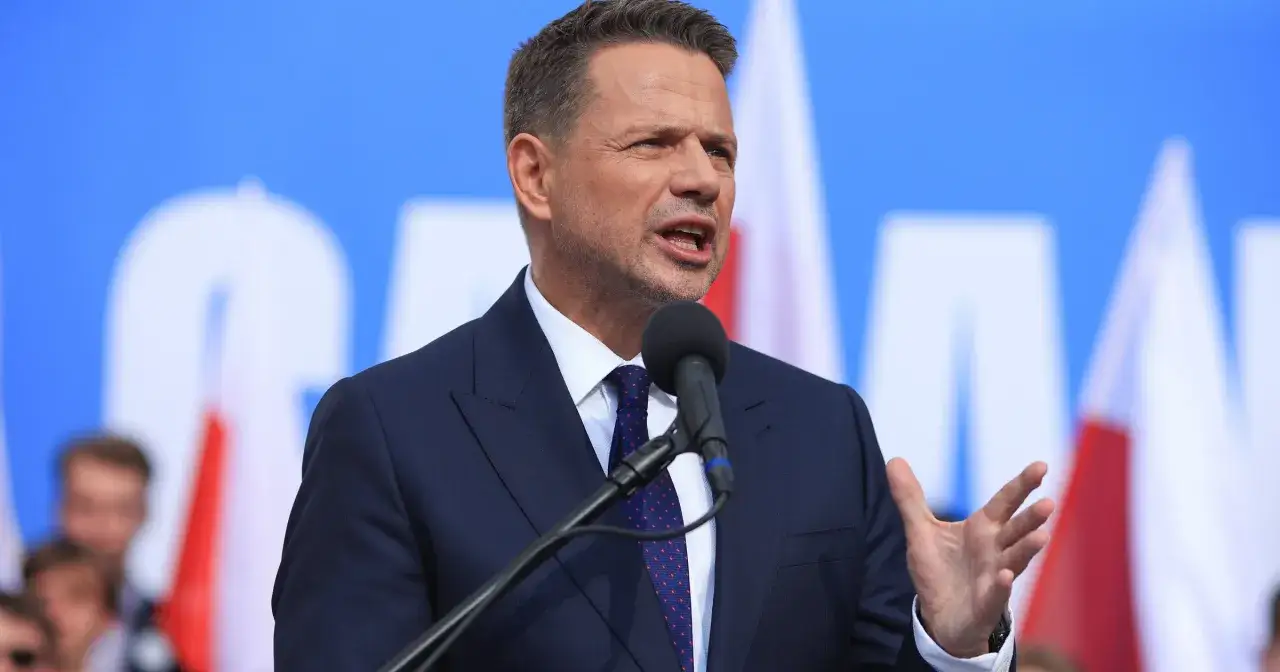 Rafał Trzaskowski, z wykształceniem, przemawia do tłumu na tle flag.