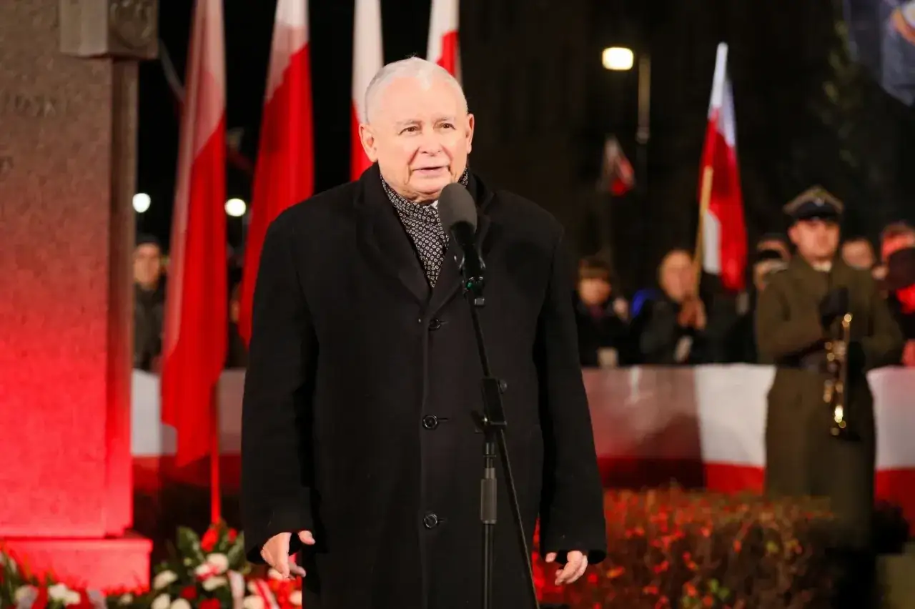 Jarosław Kaczyński przemawia na tle polskich flag, symbolizując suwerenną Polskę. W tle widać żołnierza z trąbką.
