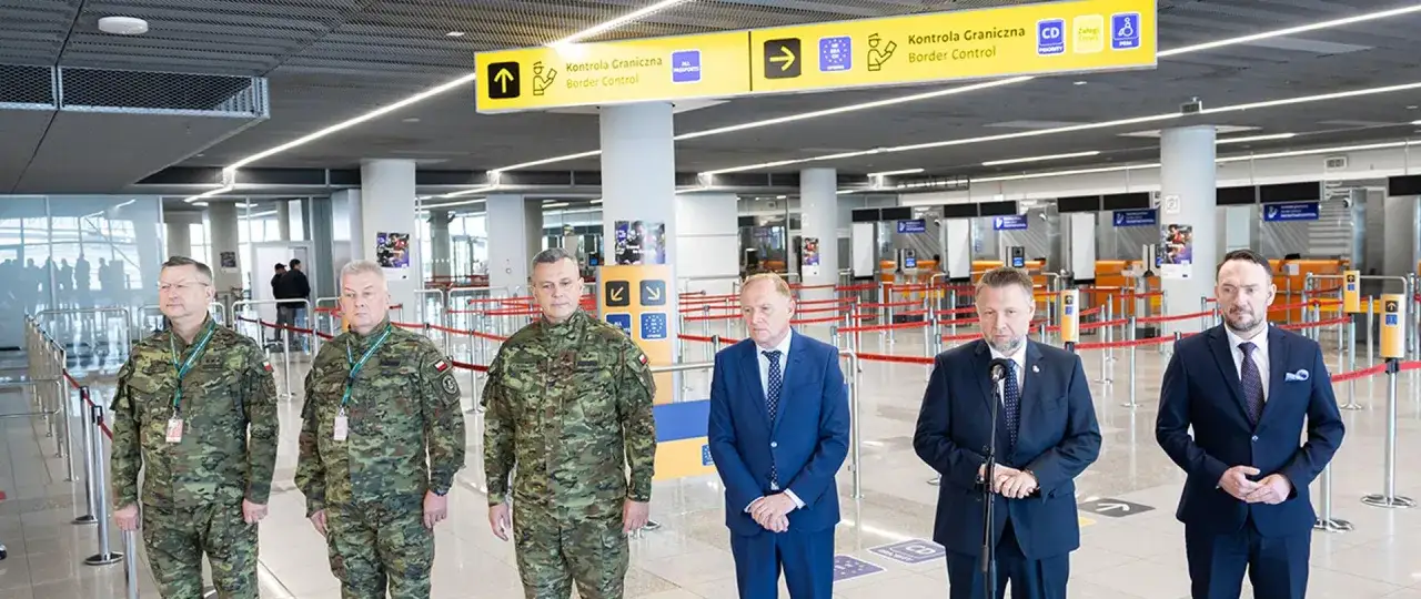 Pięciu mężczyzn, czterech w mundurach wojskowych i dwóch w garniturach, stoi przed stanowiskami kontroli granicznej.