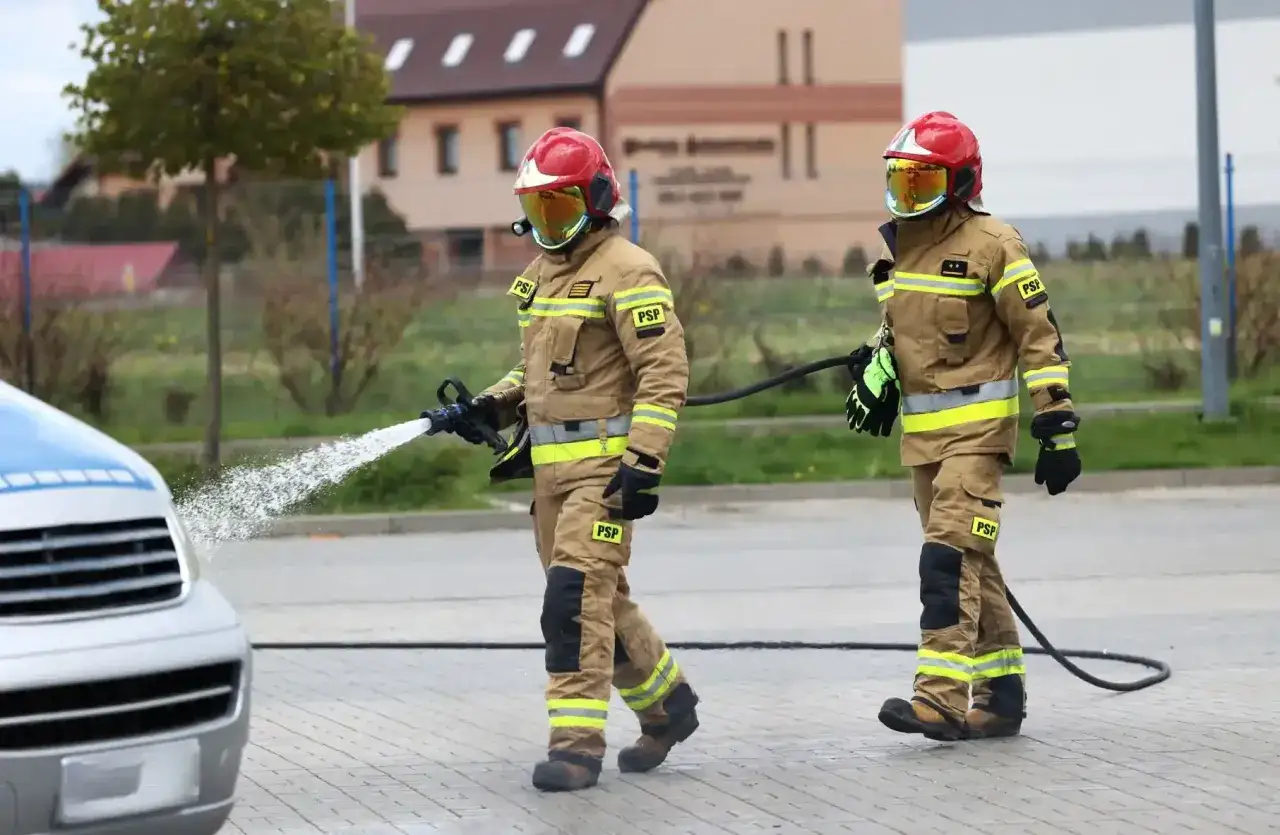 Dwóch strażaków w akcji. Jeden z nich polewa wodą samochód, drugi trzyma wąż. Ciekawe, ile zarabia strażak za taką pracę.